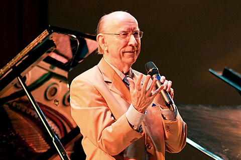 An older gentleman in a tan suit is speaking on stage with a microphone in his hand with a piano in the background.