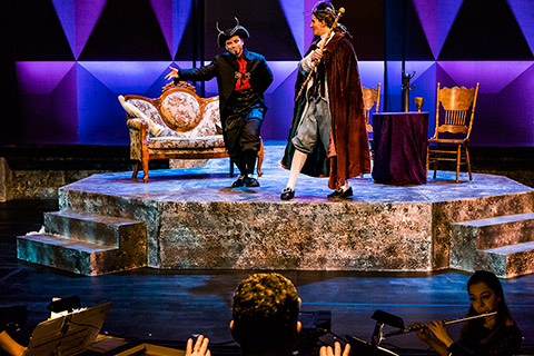 Two comically dressed men perform on stage with some furniture on it, in the foreground there is a live orchestra playing.